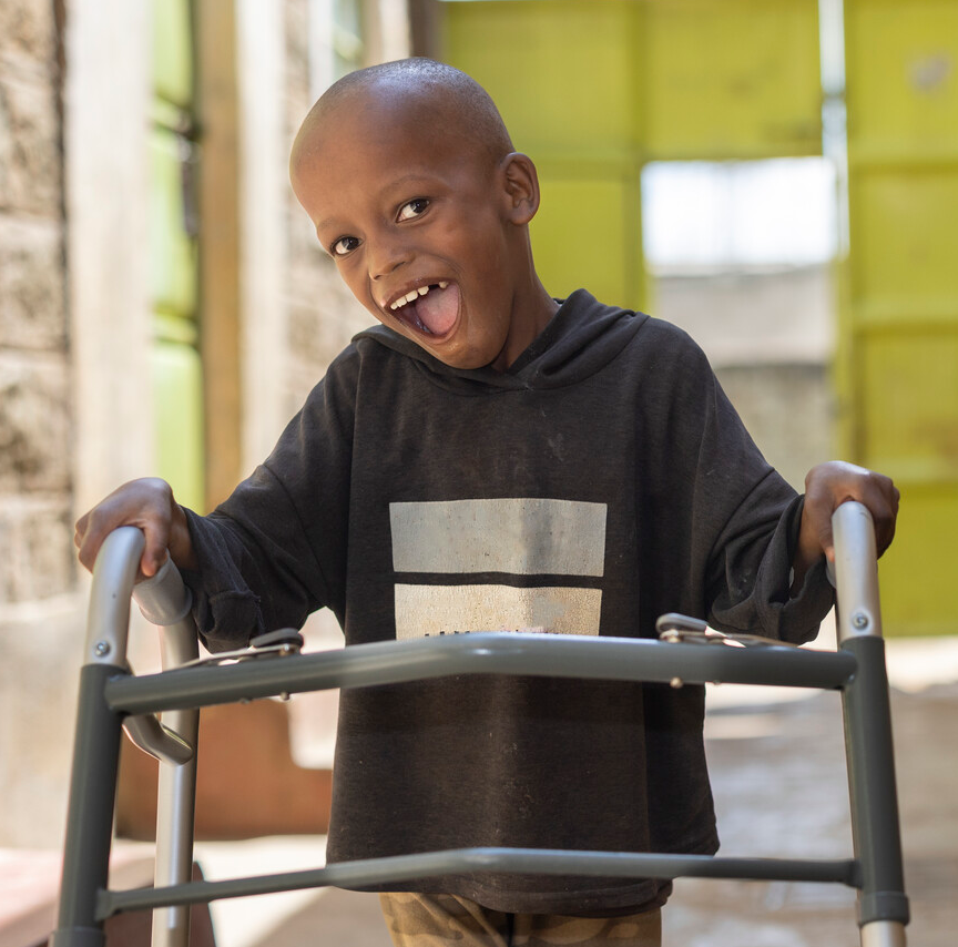 A smiling young boy in a black hoodie uses a walker outdoors, expressing joy and confidence.