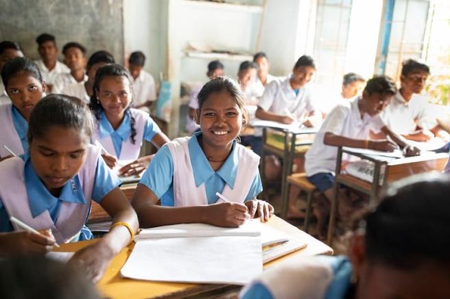 A smiling schoolgirl in a blue and white uniform writes in her notebook, sitting in a classroom with other students.