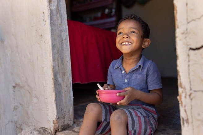 A happy young boy in a blue shirt and striped shorts sits in a doorway, holding a pink bowl with a spoon, smiling as he looks up.
