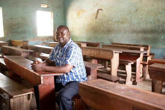 A man wearing a blue plaid shirt sits at a wooden desk in a classroom with empty wooden benches and a weathered interior.